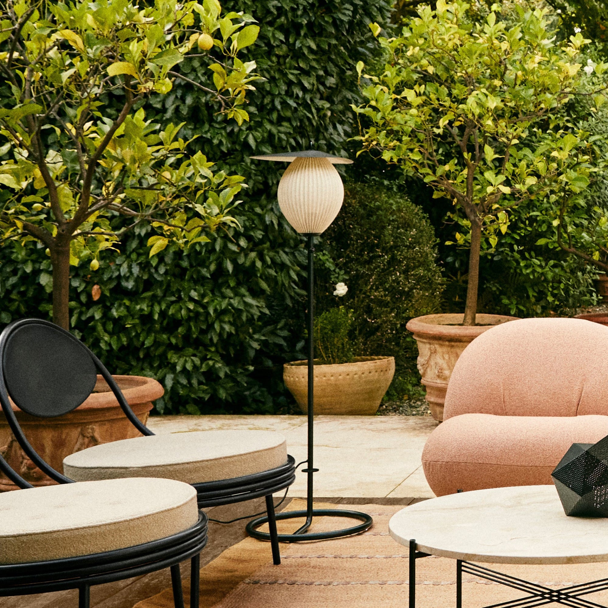 A slim black floor lamp with a ribbed cream globe shade, on a patio among potted lemon trees