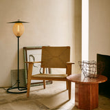 A black floor lamp with a round glowing amber shade beside a woven wooden armchair and a red stone side table