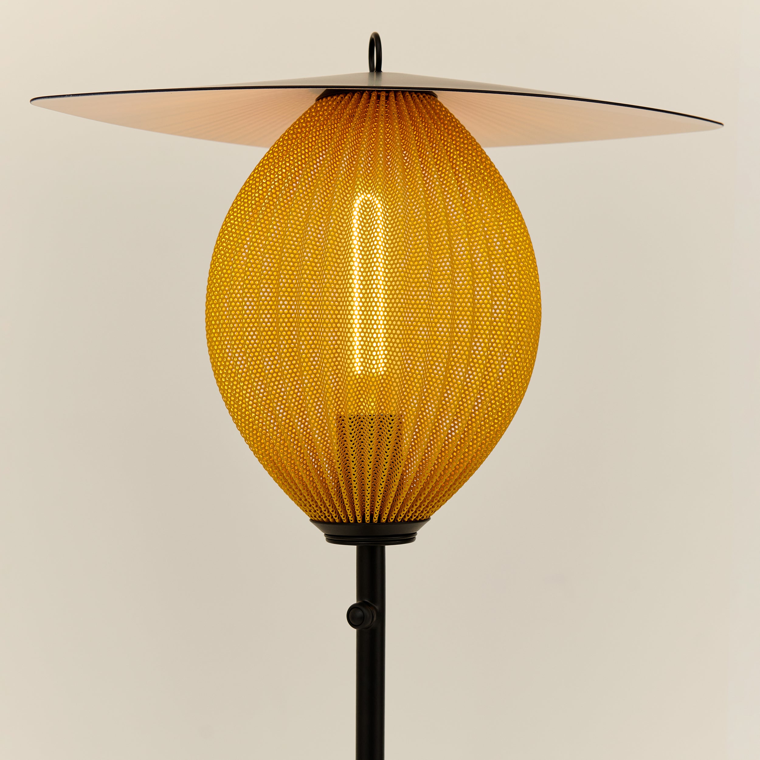 A floor lamp with a glowing yellow perforated oval shade, a wide flat disc top and a black stem