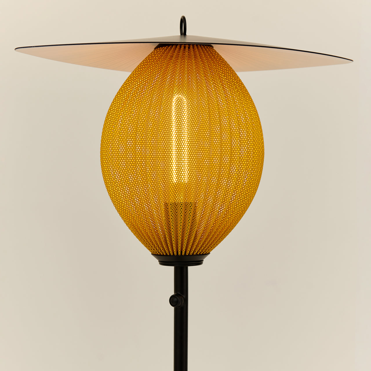 A floor lamp with a glowing yellow perforated oval shade, a wide flat disc top and a black stem