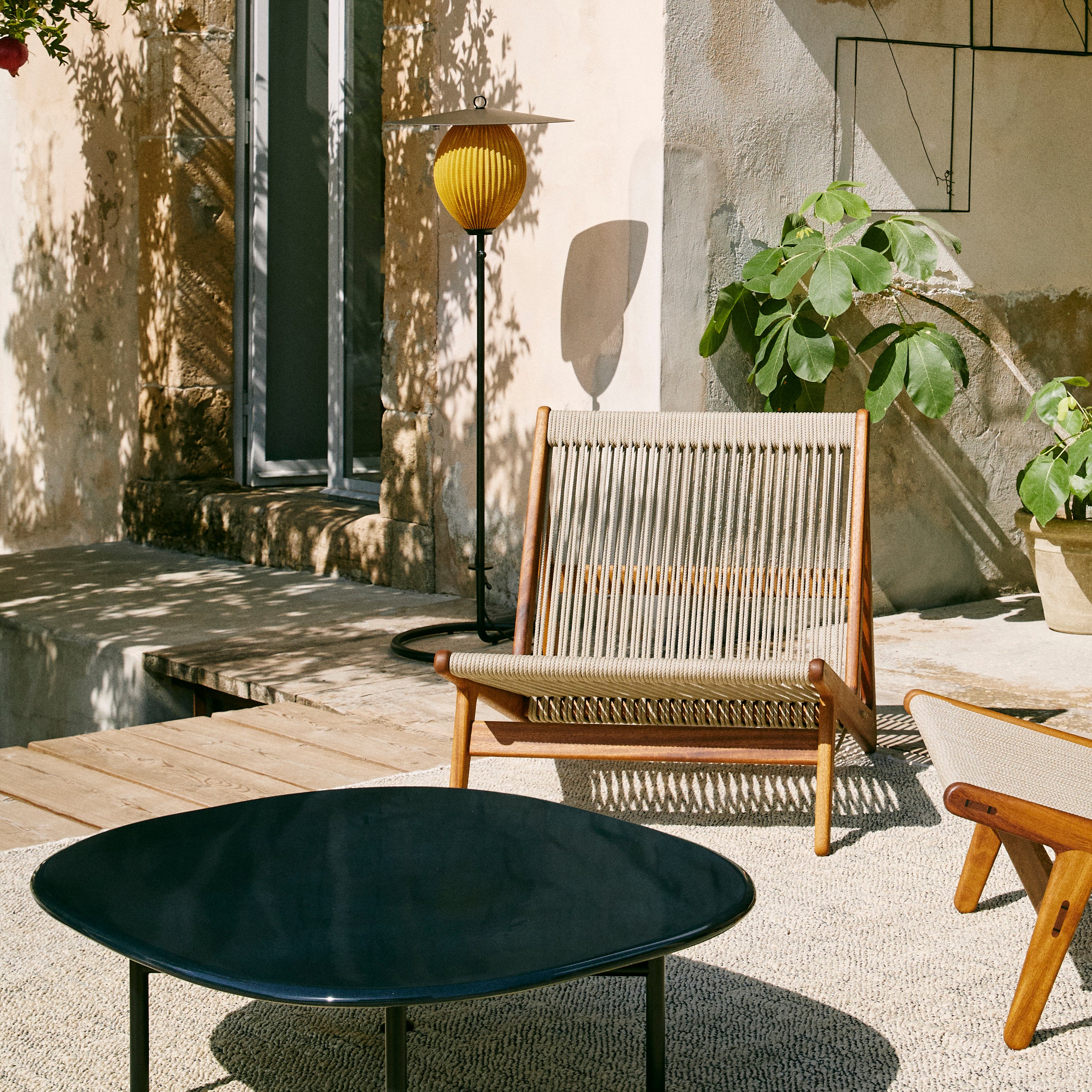 A tall black floor lamp with a yellow ribbed shade beside a rope-seat wooden chair on a sunlit terrace