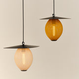 Two glowing pendant lights with wide black disc tops and perforated egg-shaped shades, one pale cream, one amber
