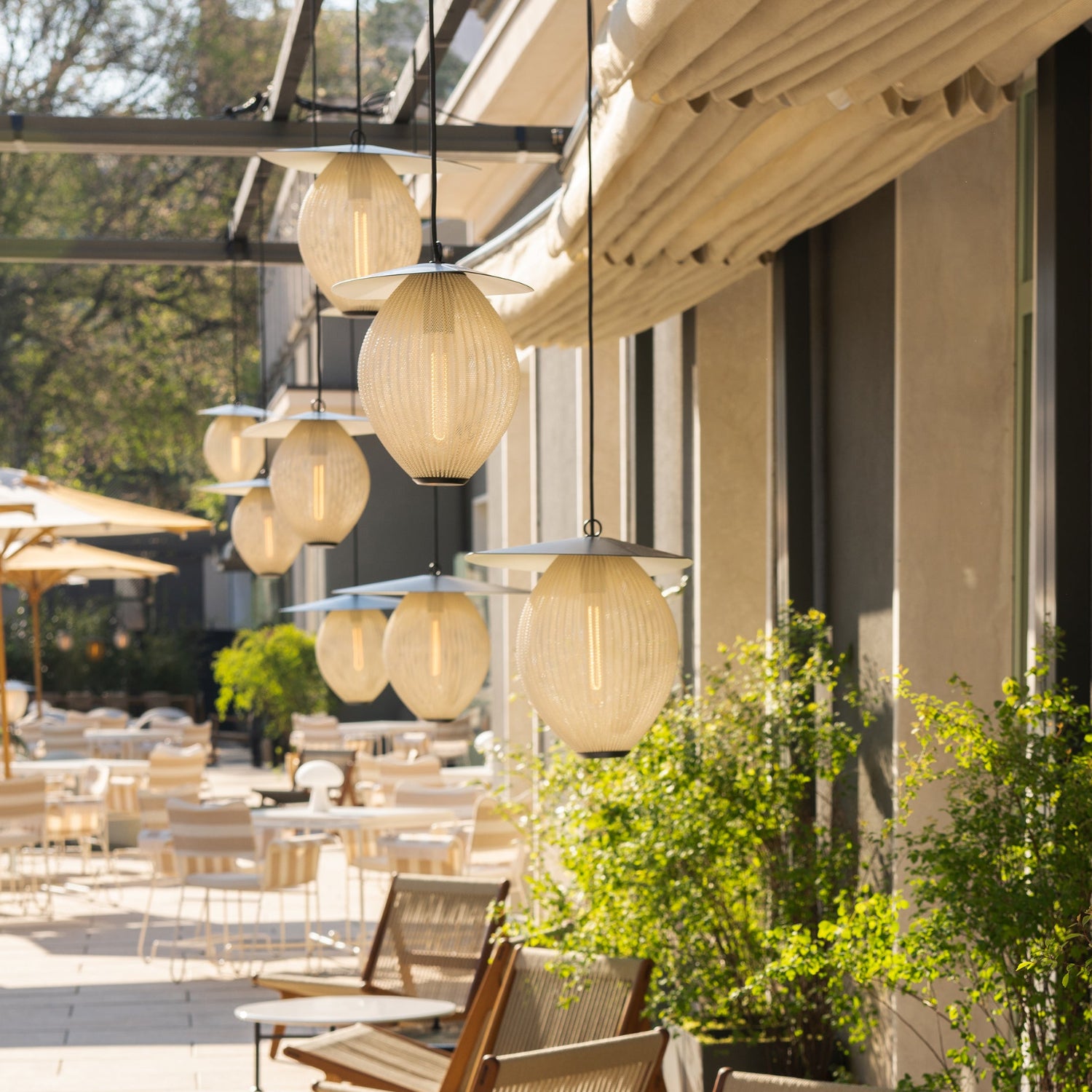 Rows of ribbed cream pendant lamps with flat grey tops hang over a sunlit outdoor café terrace