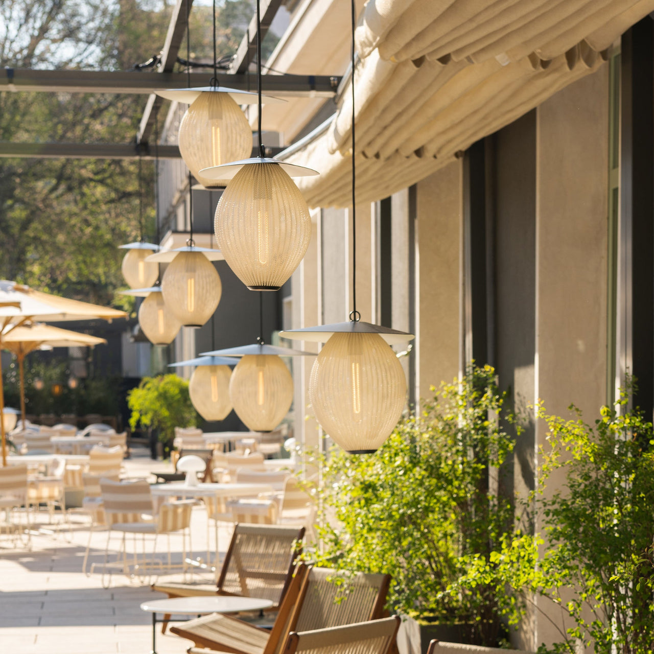 Rows of ribbed cream pendant lamps with flat grey tops hang over a sunlit outdoor café terrace