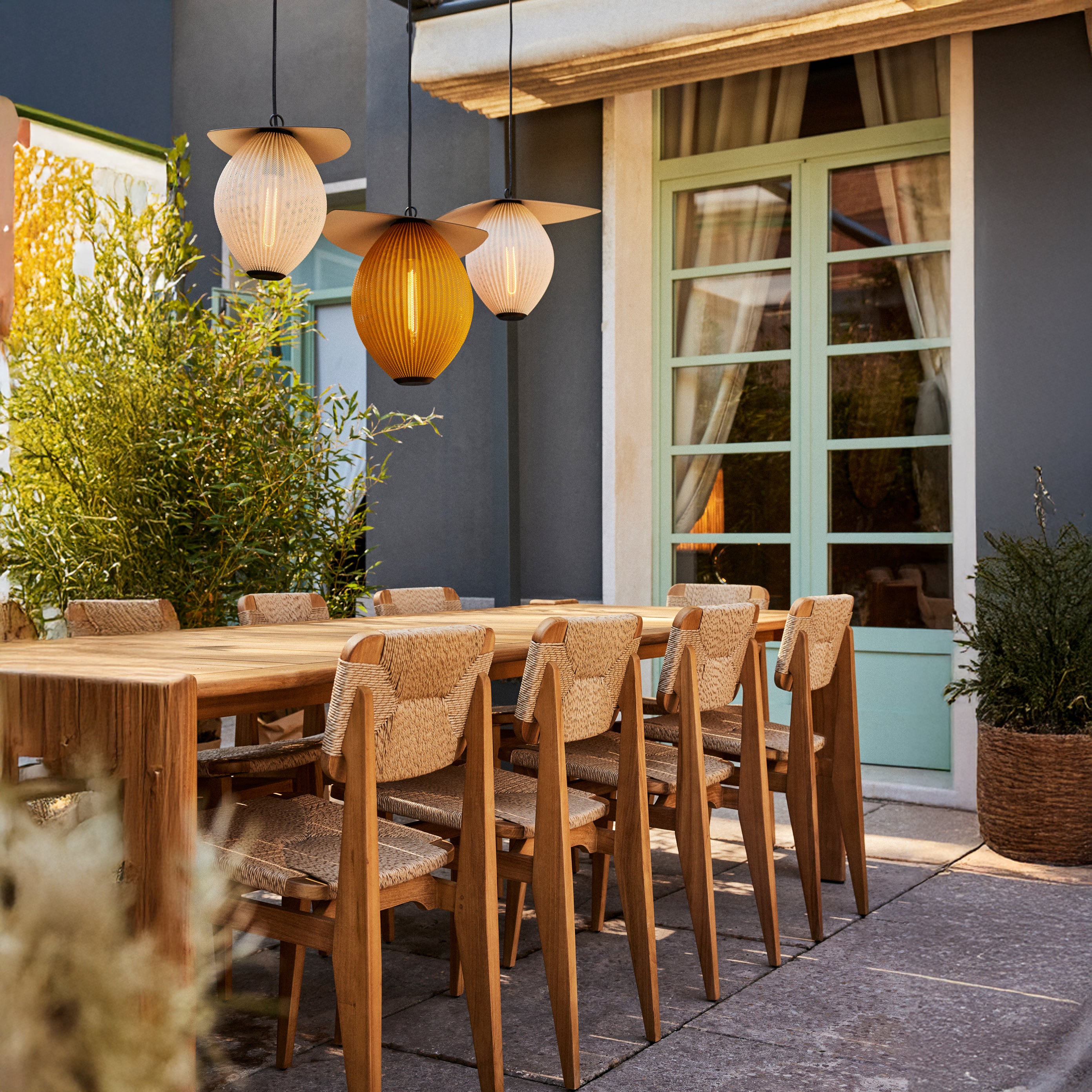 Three pleated pendant lights, one amber, above a wooden table with woven-seat chairs on a patio
