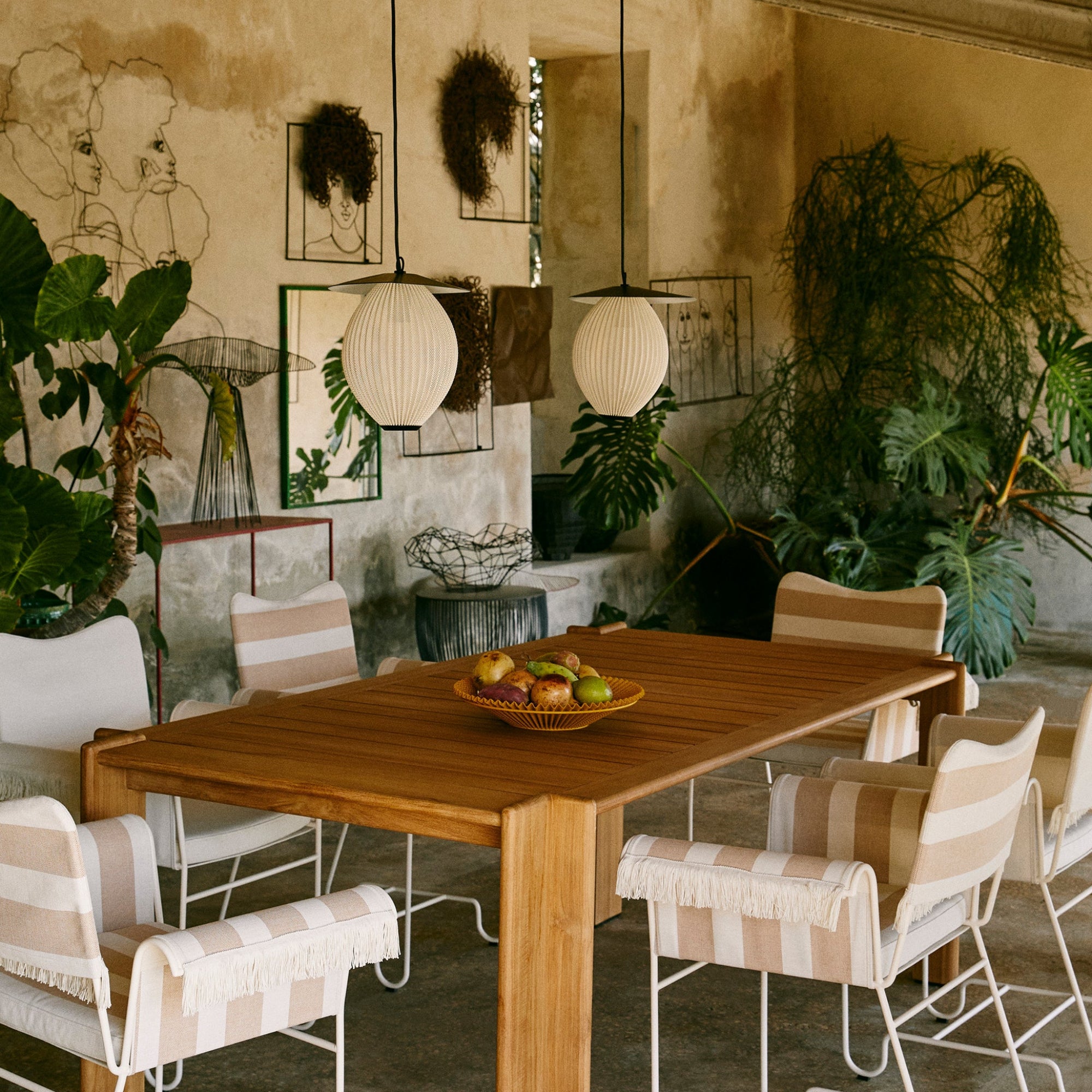 Two pale ribbed globe pendants hang over a teak table with striped chairs in a plant-filled courtyard