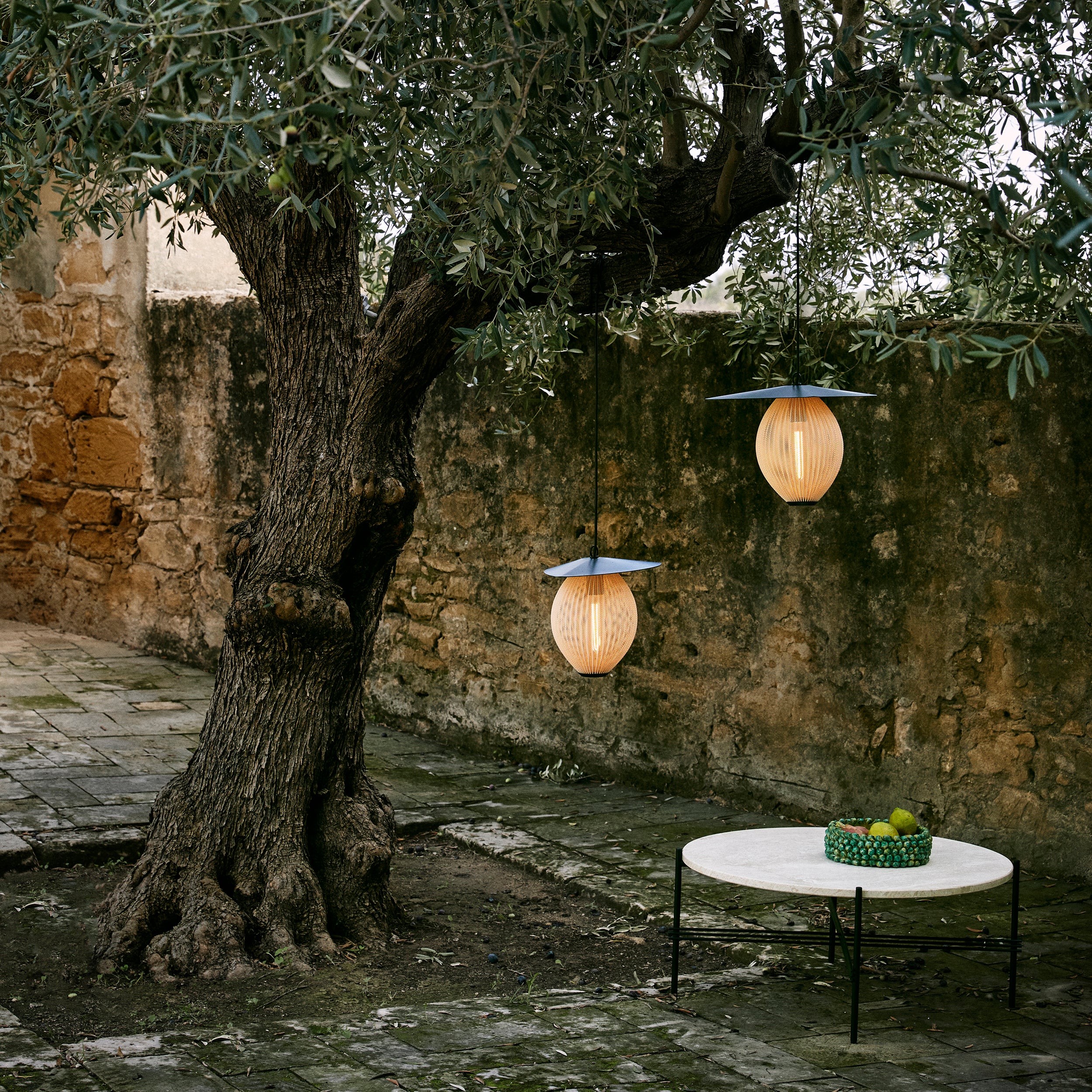 Two glowing ribbed pendant lamps with flat dark shades hang from an olive tree in a stone courtyard