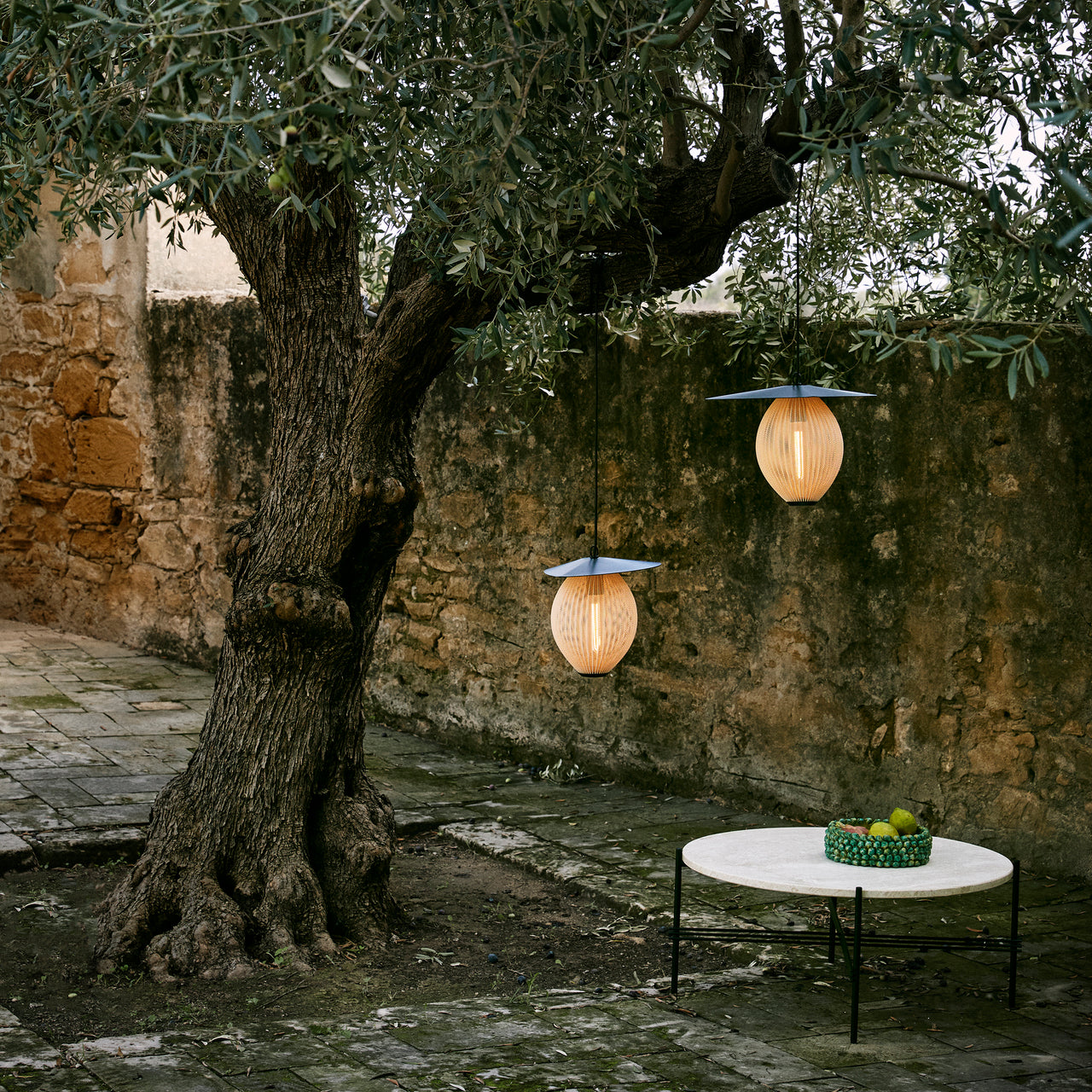 Two glowing ribbed pendant lamps with flat dark shades hang from an olive tree in a stone courtyard