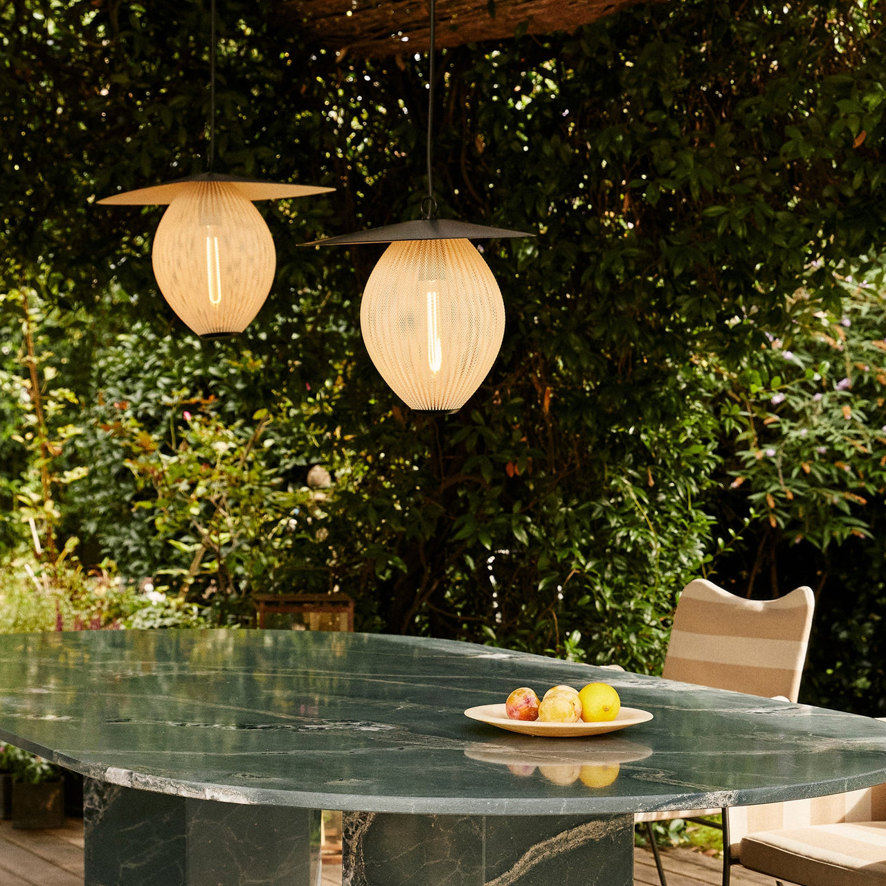 Two glowing woven pendant lamps with flat dark shades hang over a green marble table outdoors