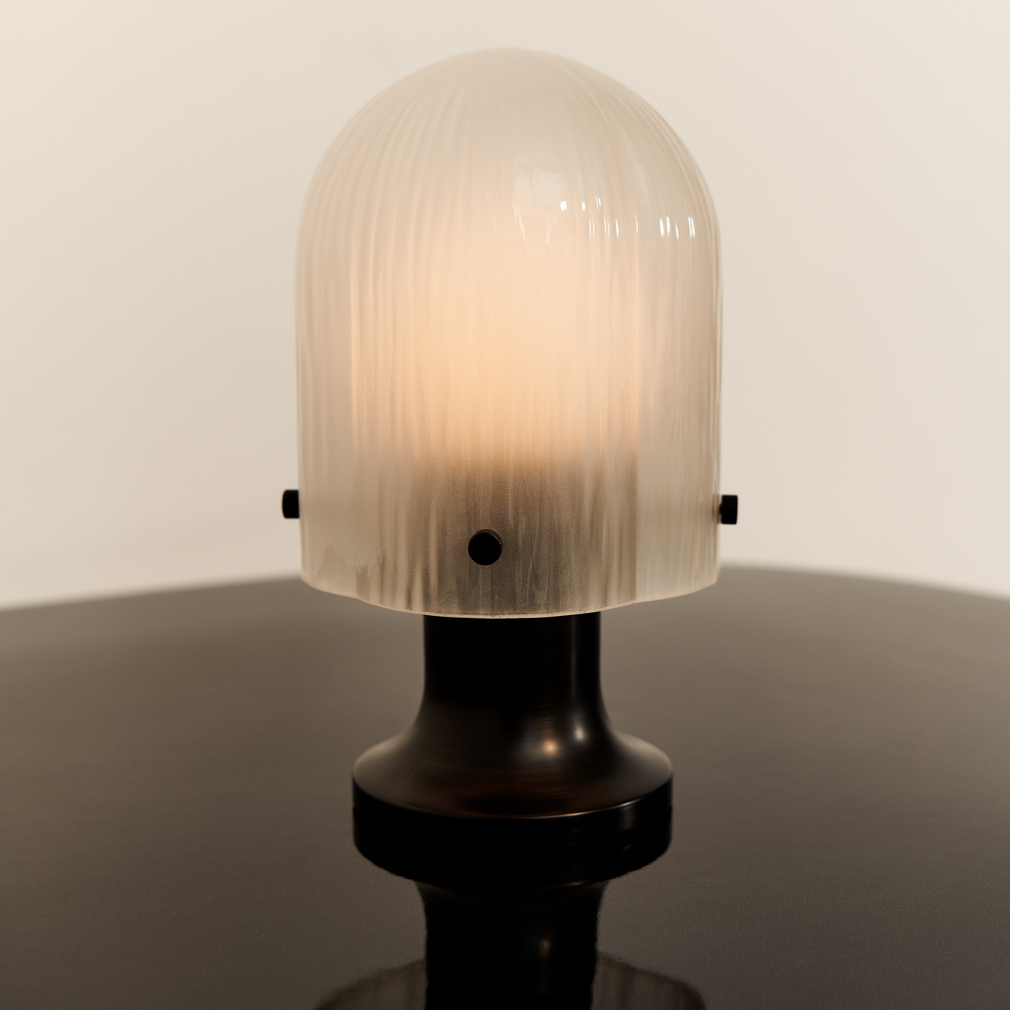 A glowing dome-shaped lamp with ribbed frosted glass on a dark bronze base, on a dark glossy surface