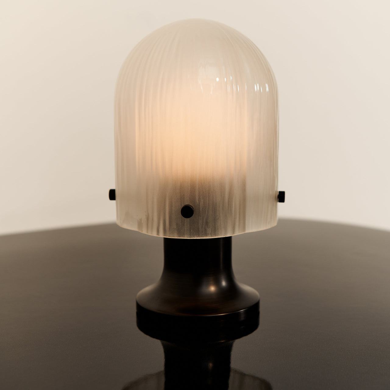A glowing dome-shaped lamp with ribbed frosted glass on a dark bronze base, on a dark glossy surface
