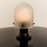 A glowing dome-shaped lamp with ribbed frosted glass on a dark bronze base, on a dark glossy surface