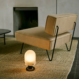 A glowing portable lamp with a ribbed frosted glass dome and dark base on a rug beside a tan chair