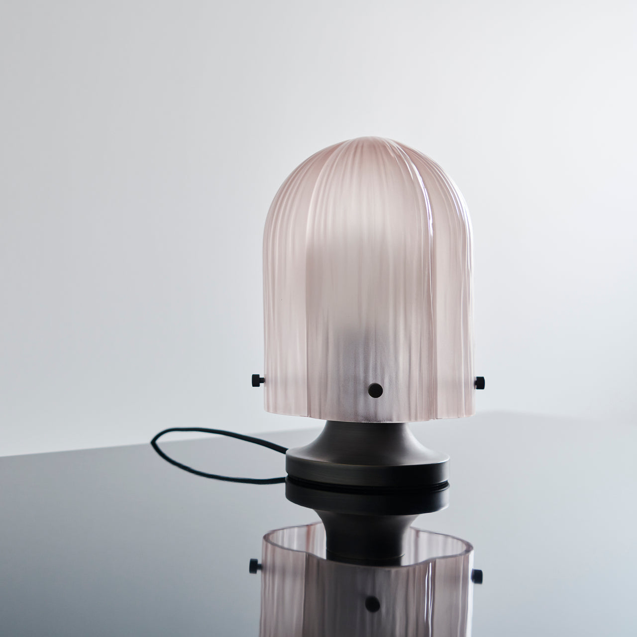 A table lamp with a ribbed pale pink glass dome on a dark metal base, reflected in a glossy surface