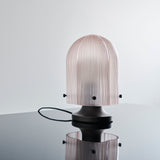 A table lamp with a ribbed pale pink glass dome on a dark metal base, reflected in a glossy surface