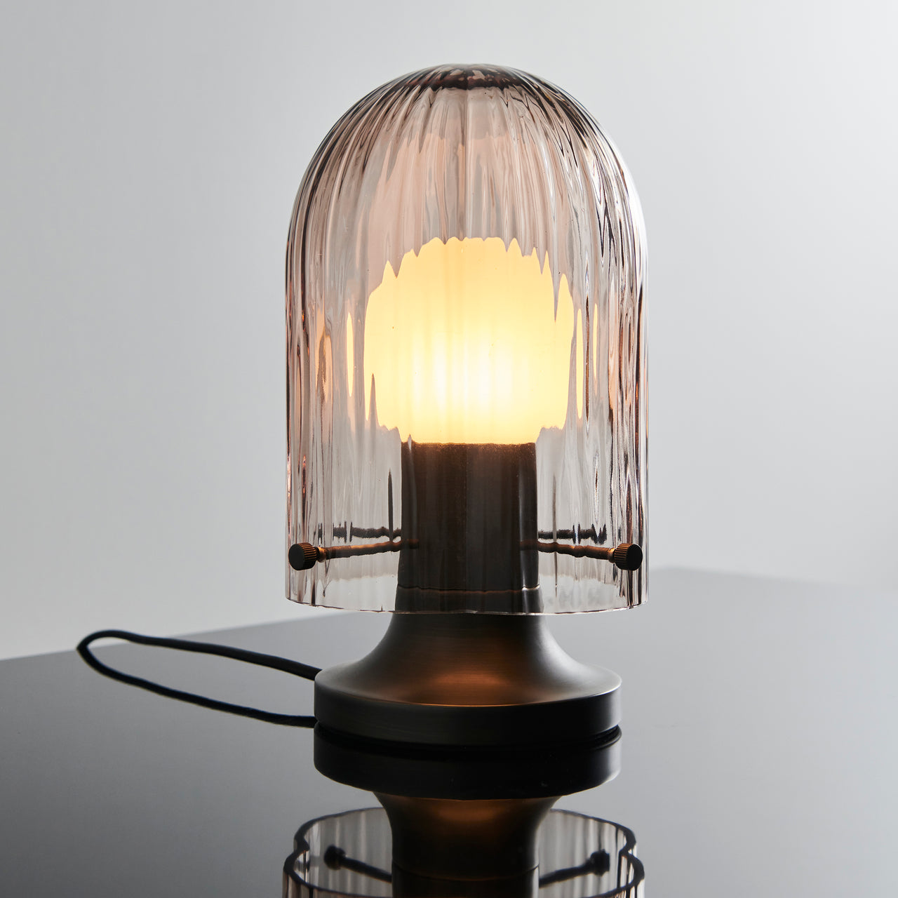 A lit table lamp with a ribbed smoky glass dome over a glowing bulb, on a dark metal base