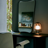 A glowing table lamp with a ribbed smoked glass dome on a dark desk beside a tall mirror