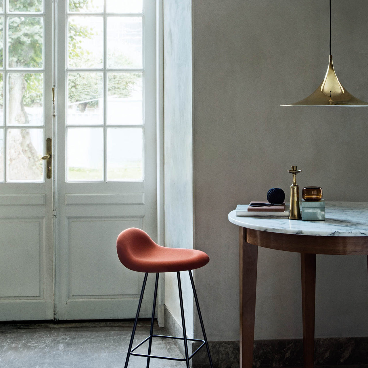 A polished brass cone-shaped pendant hangs above a marble-topped wooden table beside a rust-red stool