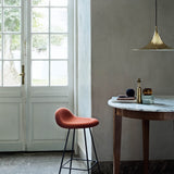 A polished brass cone-shaped pendant hangs above a marble-topped wooden table beside a rust-red stool