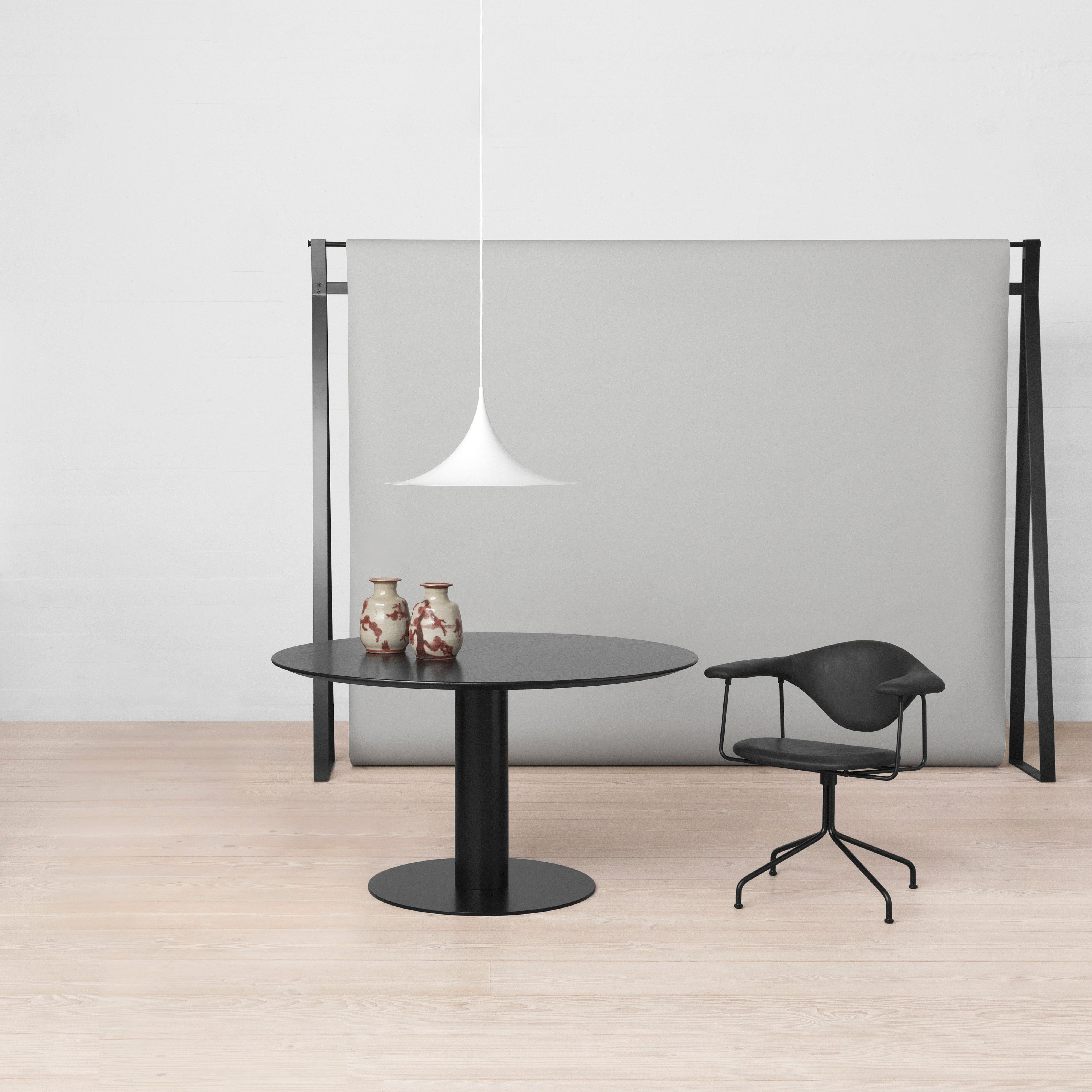 A white trumpet-shaped pendant hangs above a round black table, a black chair and a grey panel