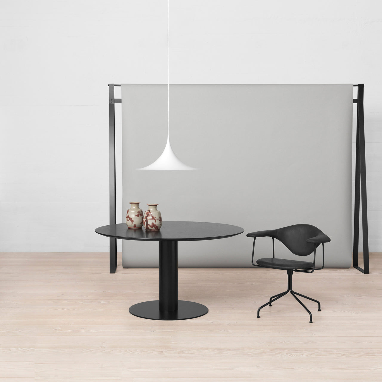 A white trumpet-shaped pendant hangs above a round black table, a black chair and a grey panel