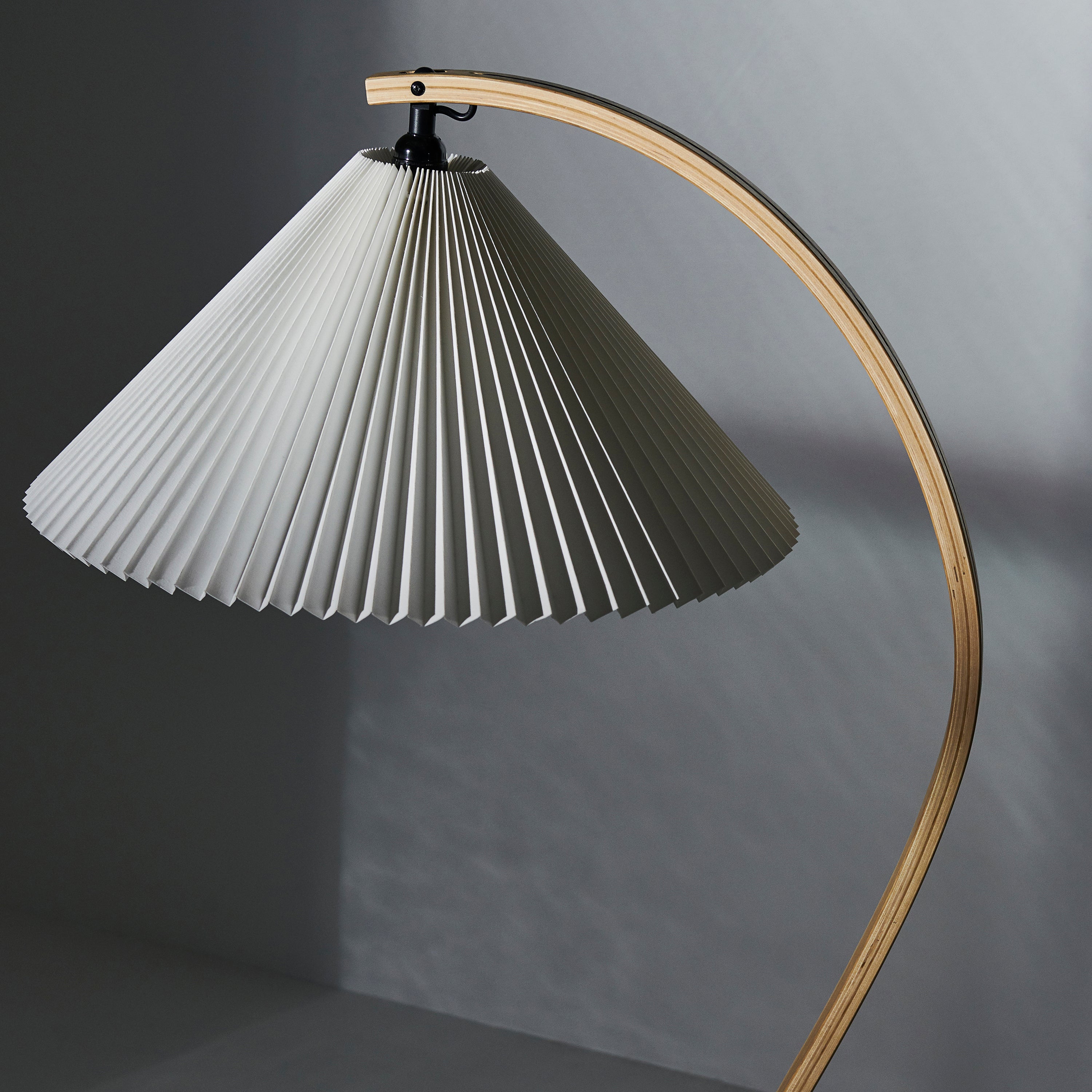 A floor lamp with a wide pleated white shade hanging from a curved light wood arm against a grey wall