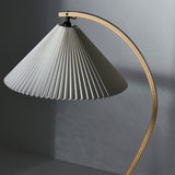 A floor lamp with a wide pleated white shade hanging from a curved light wood arm against a grey wall