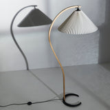 A floor lamp with a curved light wood arm, pleated white shade and black crescent base, casting a shadow on the wall