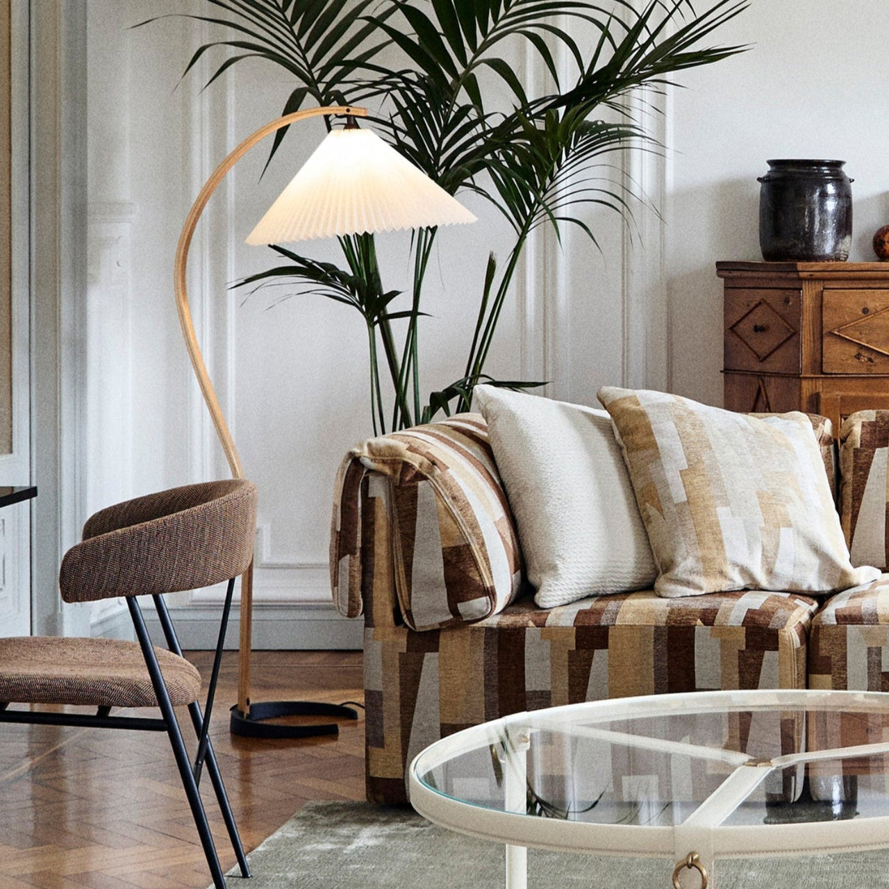 A floor lamp with a curved wooden arm and pleated white shade beside a patterned sofa and palm