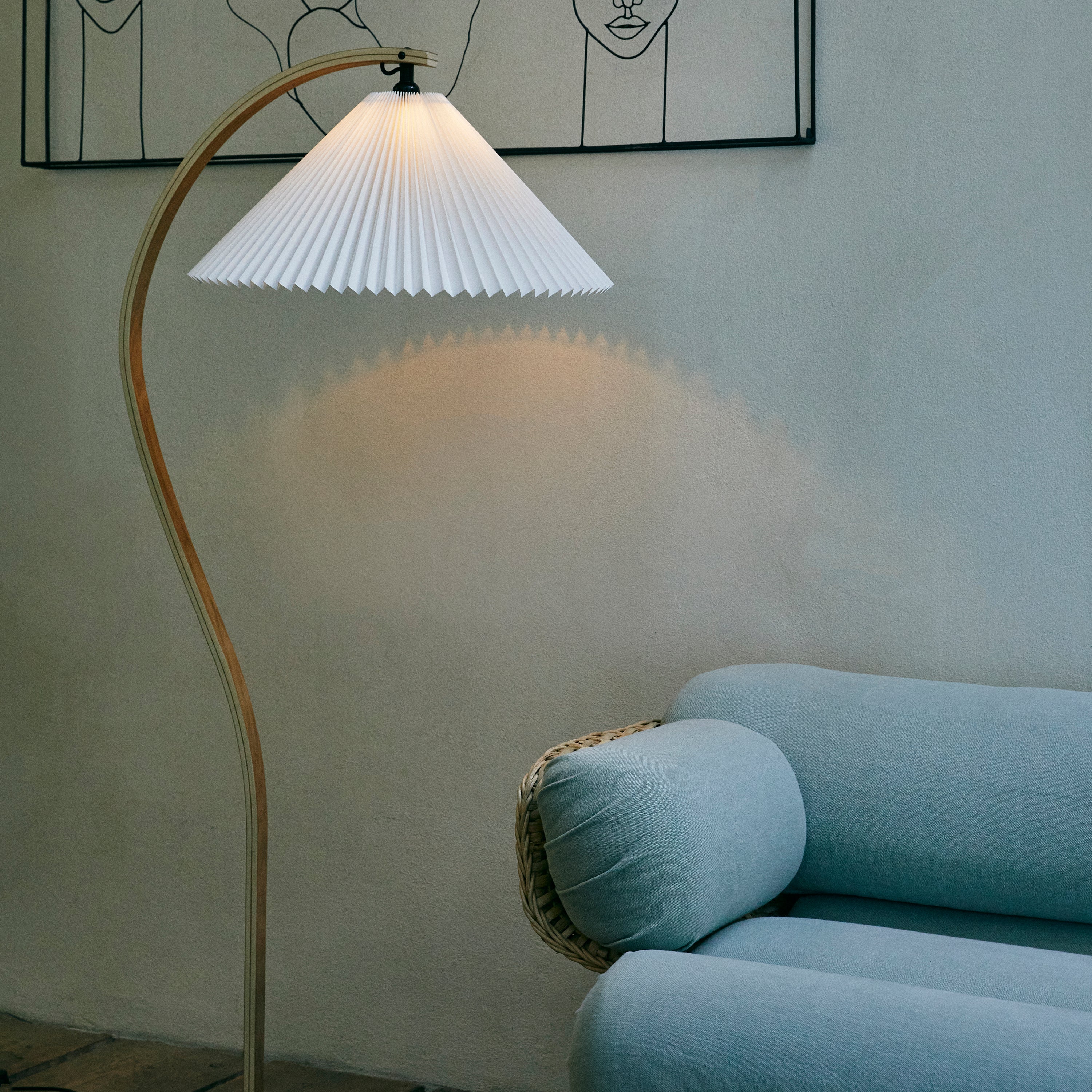 A floor lamp with a curved light wood stem and white pleated shade beside a pale blue sofa