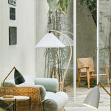 A floor lamp with a pleated white shade on a curved pale wood arm beside a rattan armchair