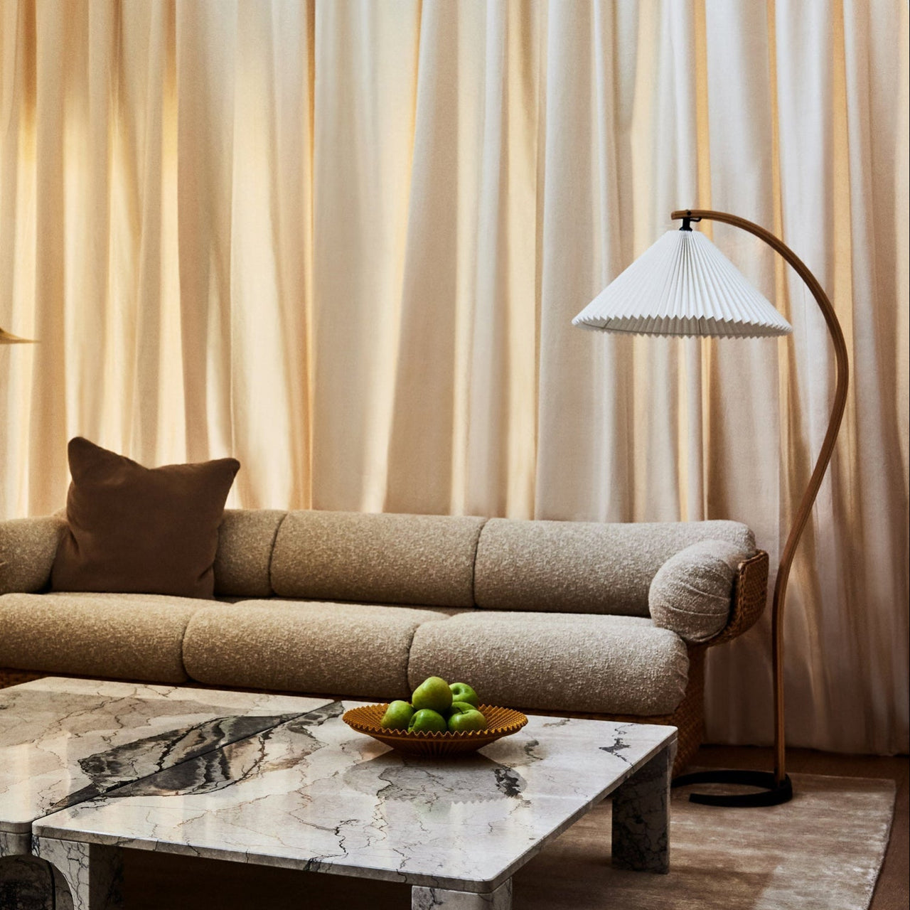 A floor lamp with a pleated white shade on a curved wooden arm beside a beige sofa and marble table