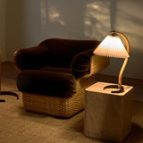 A lit table lamp with a pleated shade and curved wood arm on a stone block beside a woven armchair