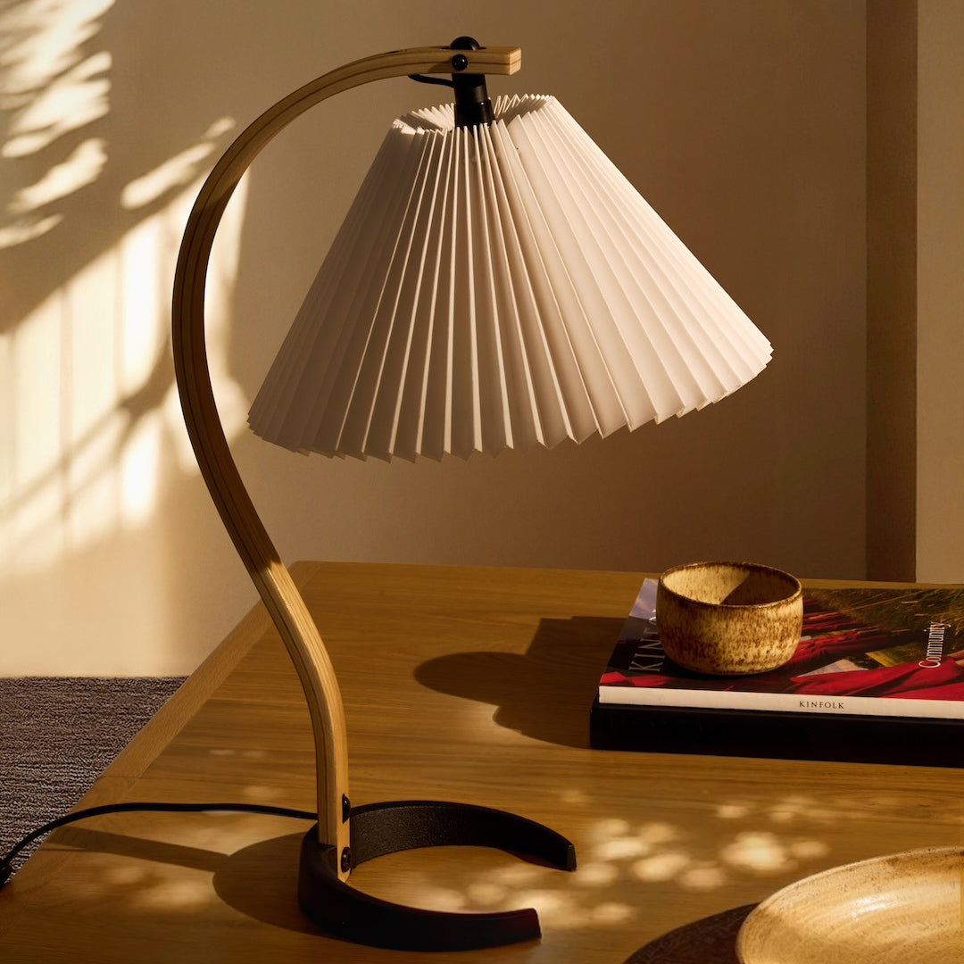 A table lamp with a curved wooden arm, black base and white pleated shade on a wooden table