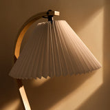Close-up of a pleated cream lampshade hanging from a curved light wood arm, against a warm sunlit wall