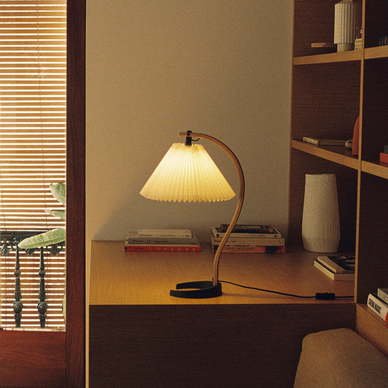 A lit table lamp with a pleated cream shade on a curved wooden arm, on a wooden desk by shelves
