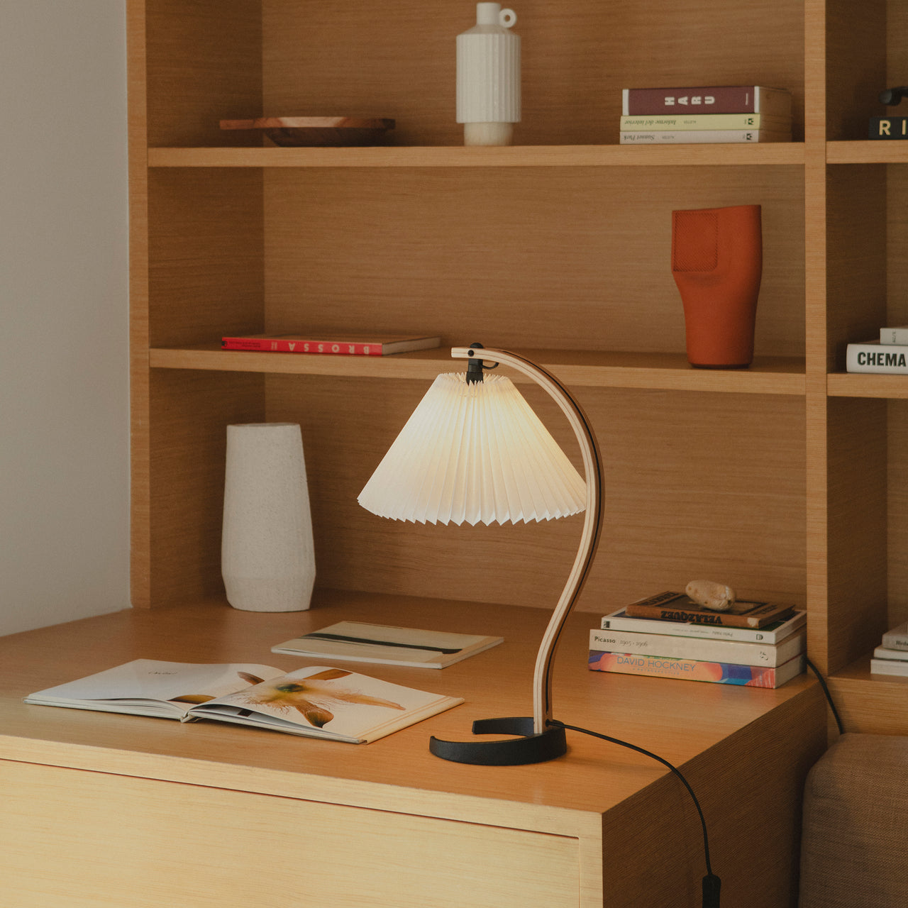 A table lamp with a pleated white shade on a curved wooden arm and black base, on an oak desk