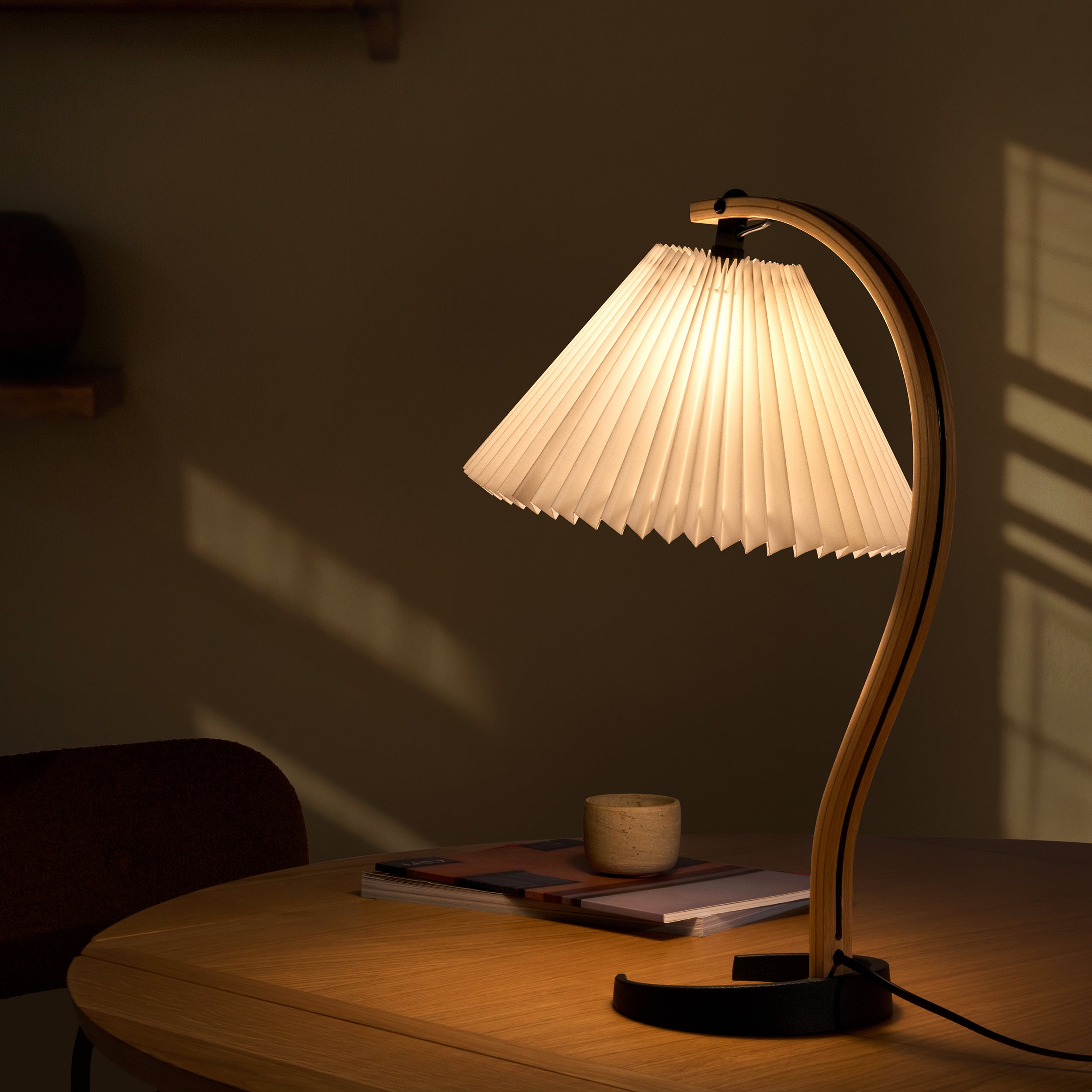 A lit table lamp with a pleated white shade and curved bentwood arm on a round oak table