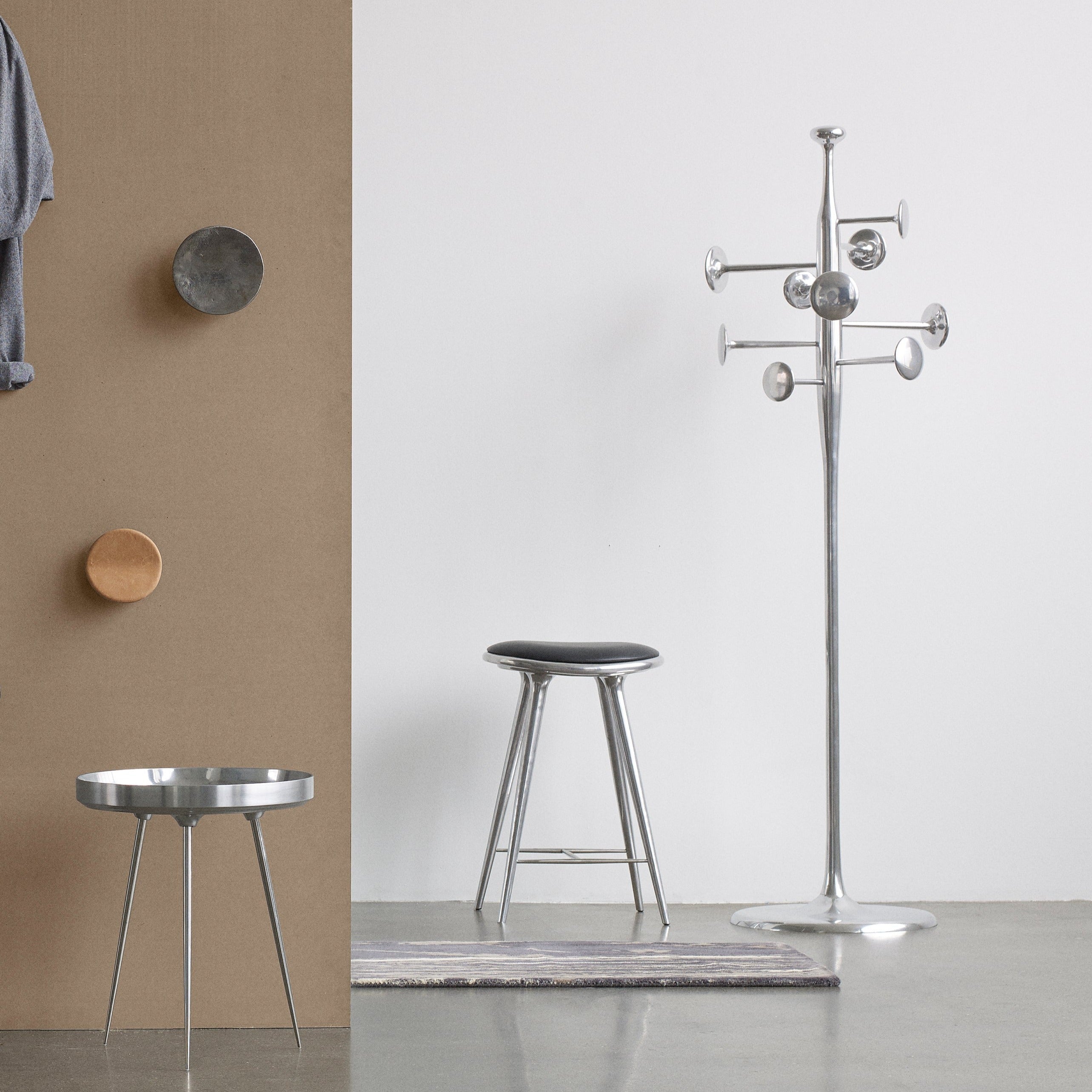 A polished aluminium coat stand with round pegs beside a metal stool and a three-legged side table