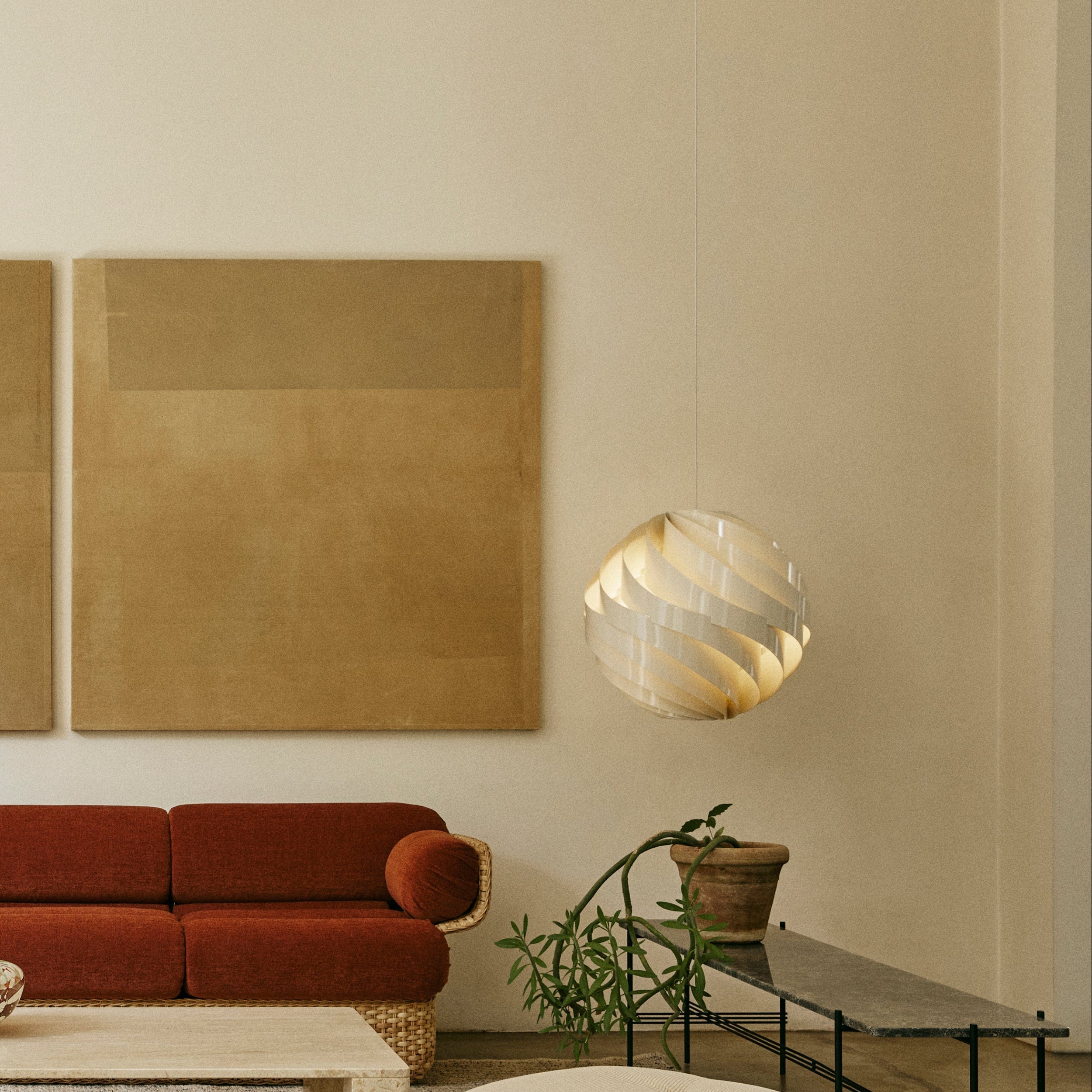 A round white pendant of twisting slats hangs beside a rust red sofa and large tan canvases