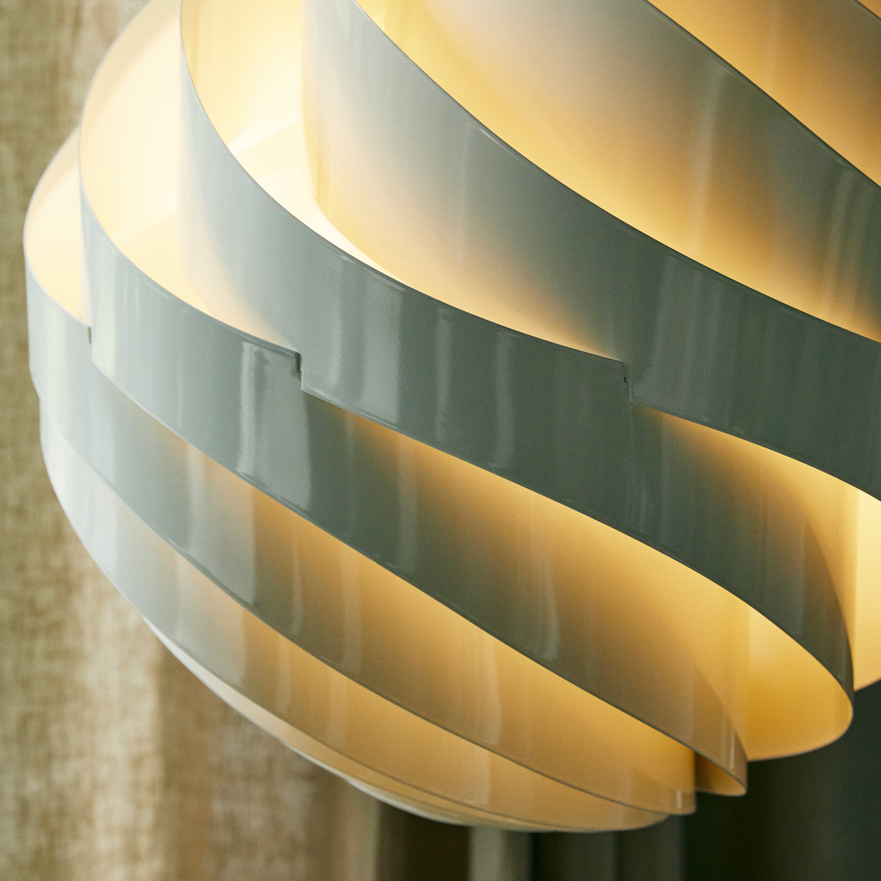 Close-up of a glowing pendant shade made of overlapping curved pale grey slats, lit warmly from within