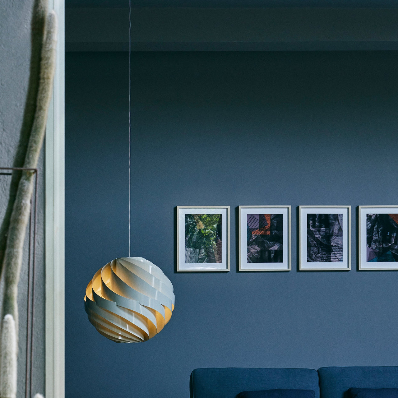 A round pendant of twisting pale slats with a warm inner glow, hung before a blue wall with framed prints