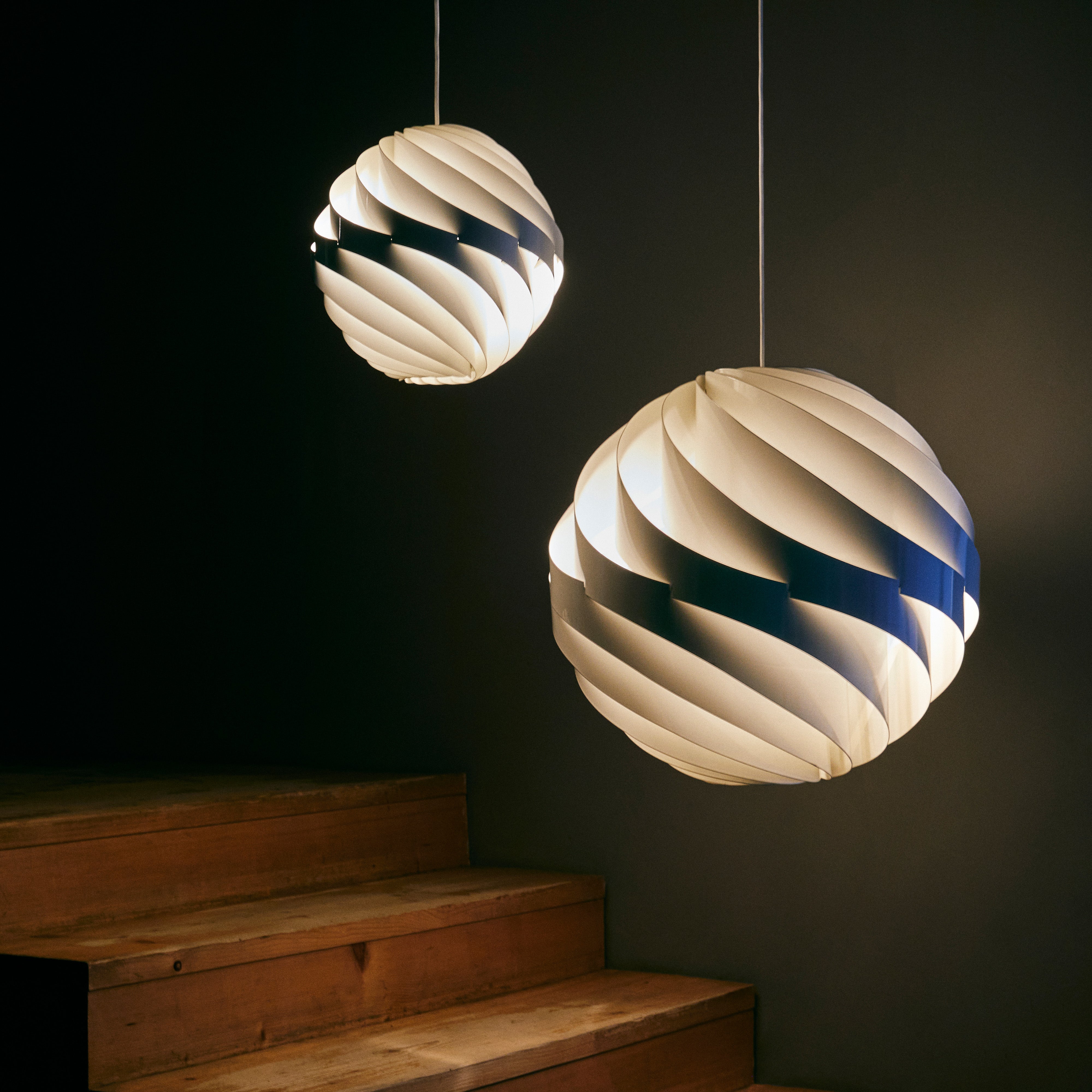 Two glowing spherical pendants made of twisting white slats, hanging beside wooden steps in a dark room