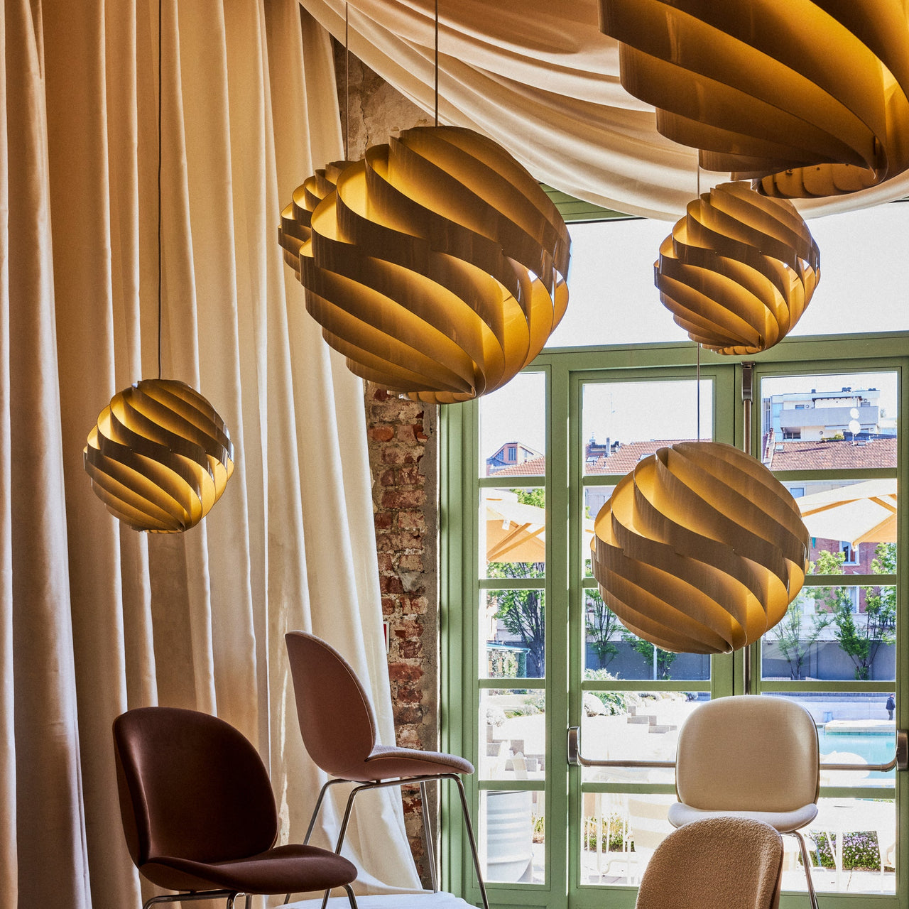Round pendants with swirled light wood slats glow near tall green windows, with cushioned chairs below