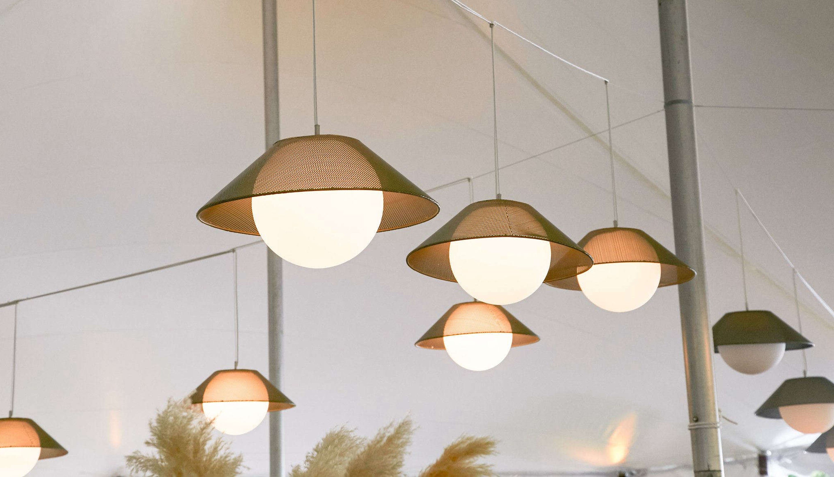 A collection of modern pendant lights with conical beige shades and glowing white globe bulbs, suspended from thin wires in a bright, airy interior space.