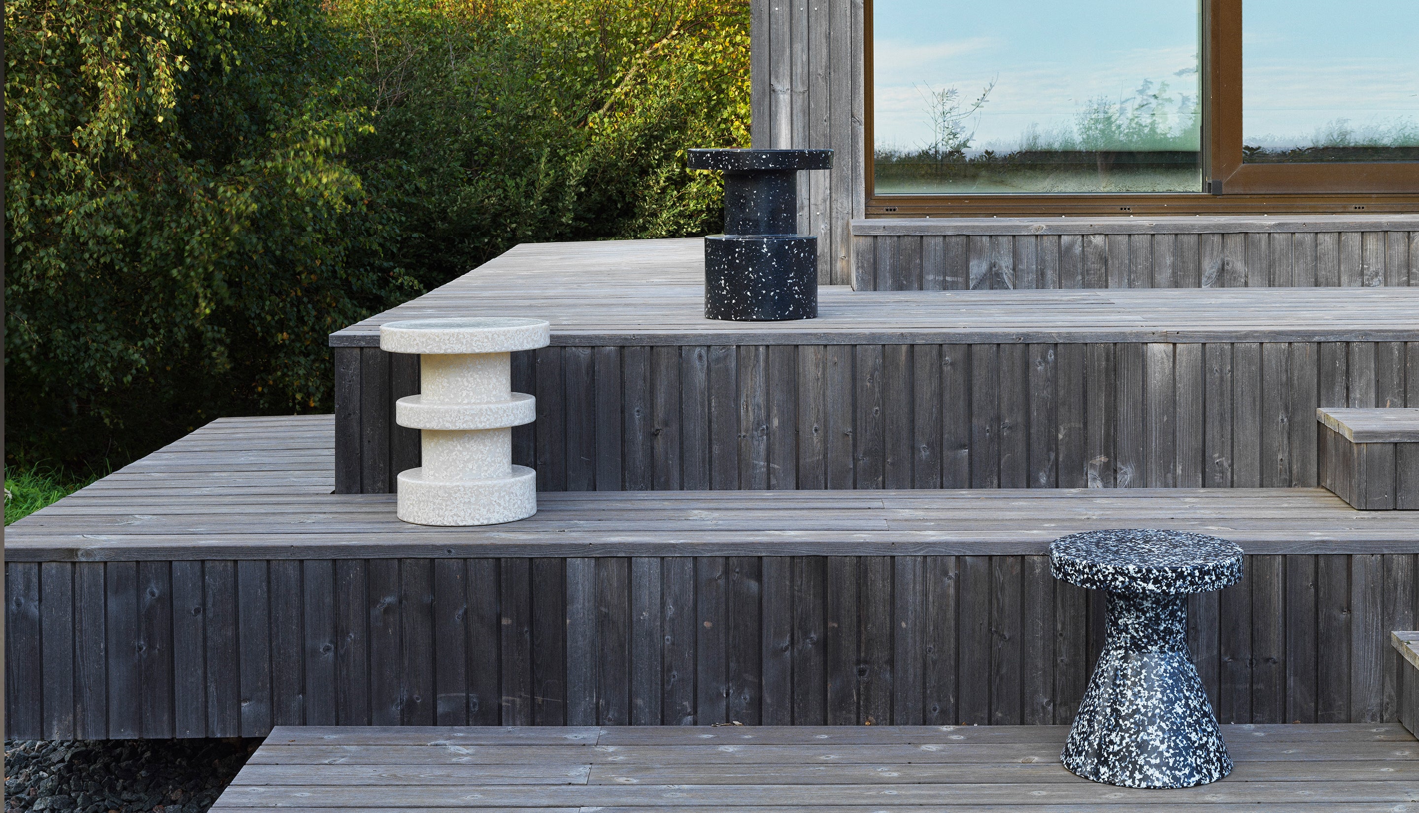 Three modern recycled stools in different textures and colors (white speckled, black speckled, and dark gray) arranged on a wooden deck outdoors.