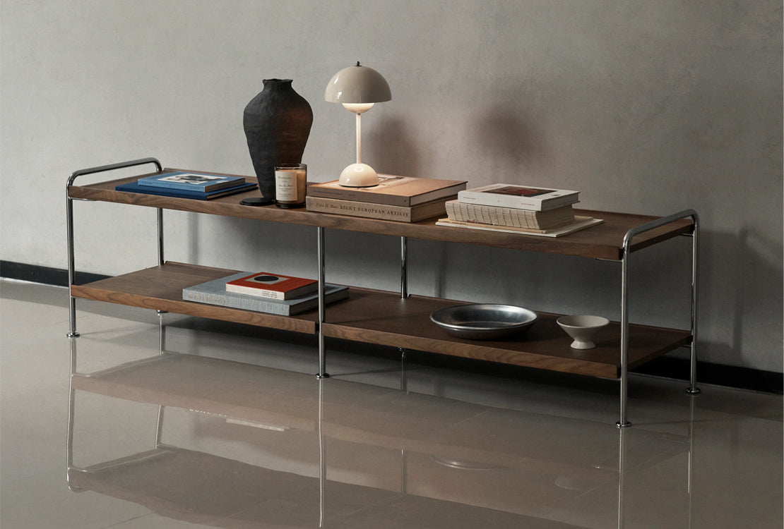 A modern two-tier wooden console shelf with a chrome metal frame, styled with books, a lamp, a ceramic vase, and decorative plates against a textured gray wall.