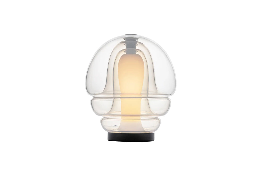 A modern table lamp with a clear glass dome shade and a black circular base, illuminated with a warm yellow light inside, set against a plain white background.