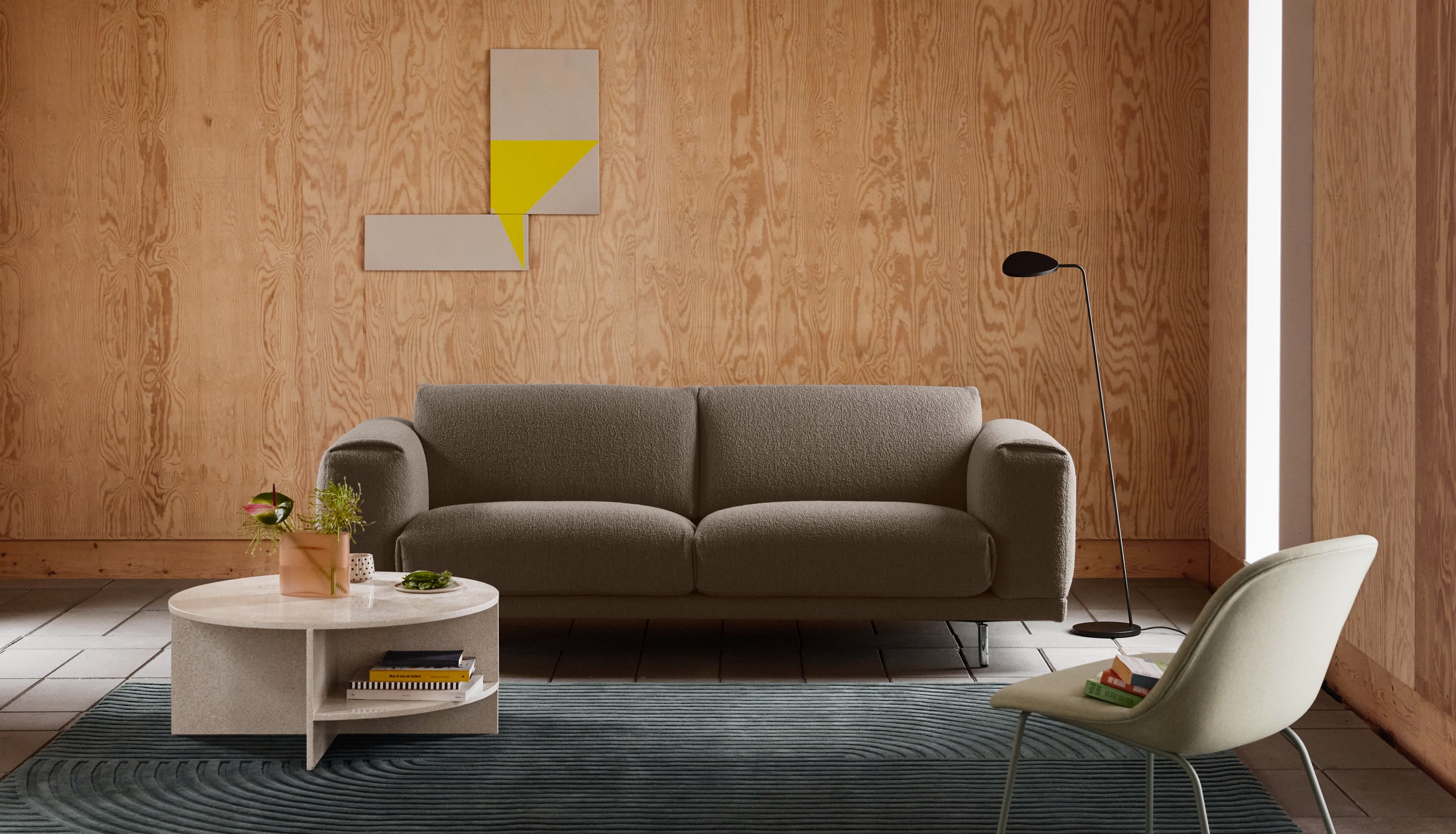 A modern living room scene featuring a gray fabric sofa as the main product, styled in a room with light wood paneling, a white coffee table, a cream chair, and a dark blue rug.