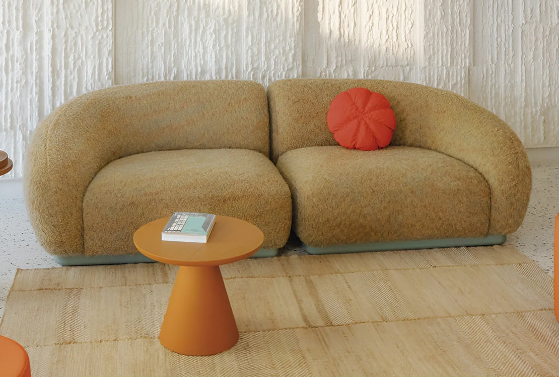 A mid-century modern curved two-seater sofa upholstered in textured beige bouclé fabric, styled with a round orange accent pillow and a small matching orange side table in a bright, modern room.